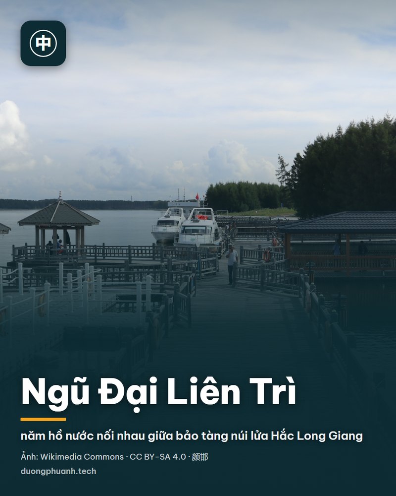 Toàn cảnh miệng núi lửa Lão Hắc Sơn nhìn từ trên vành miệng: lòng chảo rộng phủ đá bazan màu đen xám, quanh vành có lối đi bộ men theo và cây bụi xanh, phía xa là cánh đồng bằng phẳng, cánh đồng dung nham sẫm màu và mặt hồ nước sáng dưới trời xanh nhiều mây
