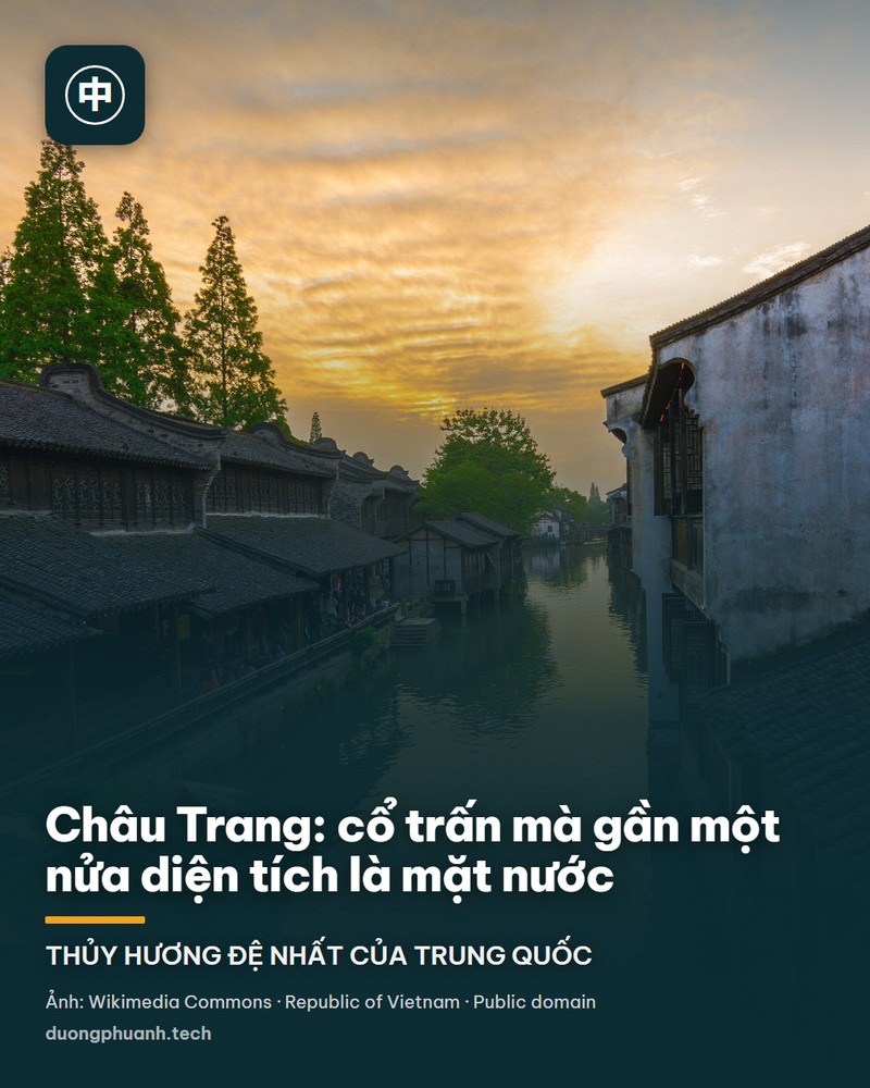 Một con kênh hẹp chạy giữa hai dãy nhà cổ tường trắng mái ngói đen của cổ trấn Châu Trang, mặt nước phản chiếu ánh vàng của bầu trời lúc hoàng hôn, hàng hiên và lan can gỗ chạy dọc bờ kênh, phía dưới bên trái là chùm lá xanh trước ống kính.
