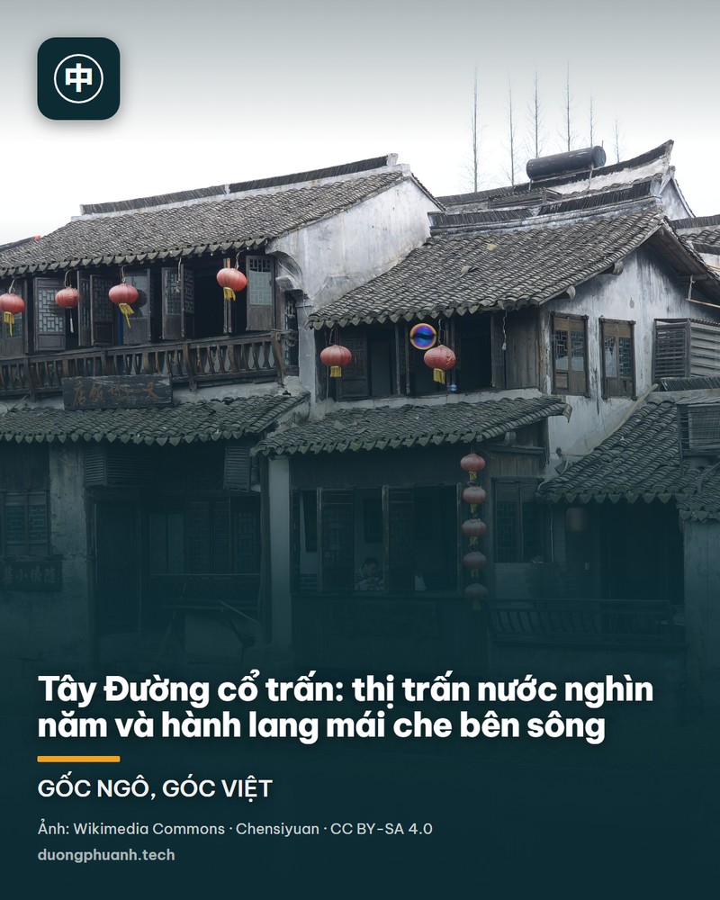Một góc cổ trấn nước Tây Đường nhìn dọc theo con kênh: hai dãy nhà cổ hai tầng mái ngói đen nằm sát mặt nước, cửa sổ chấn song gỗ, đèn lồng đỏ treo dọc hiên, cành liễu và cành cây trụi lá vắt ngang bầu trời xám nhạt, giữa kênh có một chiếc thuyền nhỏ đi về phía cây cầu đá ở xa.