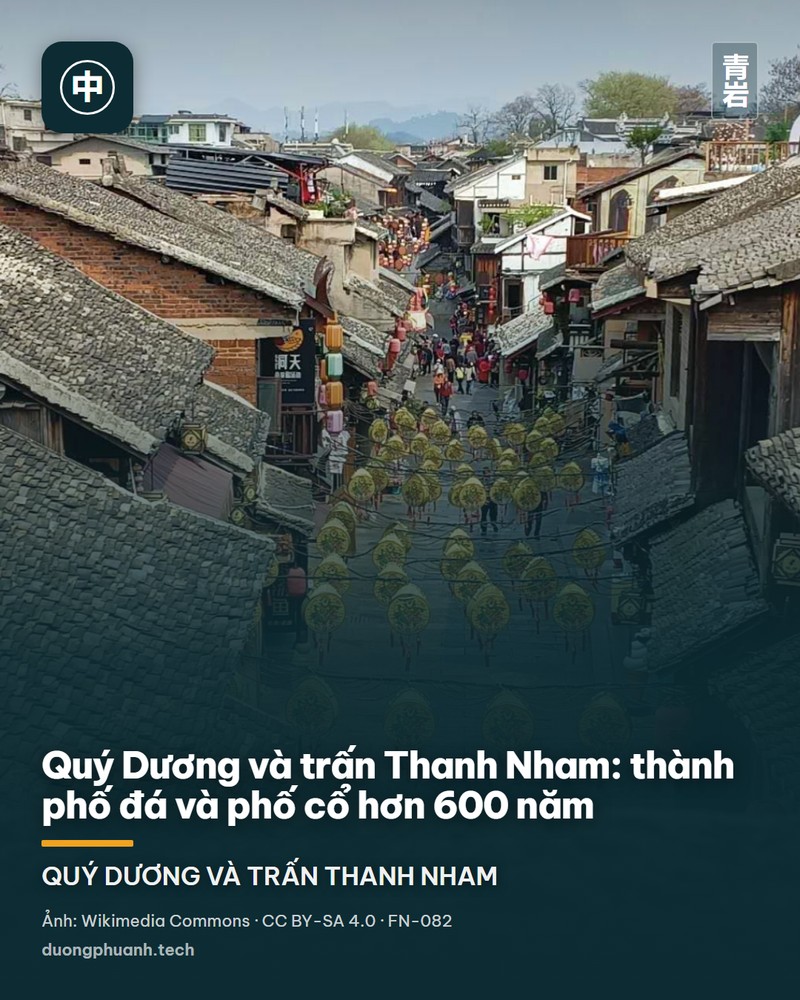 Phố chính của trấn cổ lát đá xám, hai bên là dãy nhà gỗ hai tầng mái ngói xám, hiên thấp treo đèn lồng đỏ và bày hàng lưu niệm; vài người đi bộ giữa phố, phía trên là bầu trời xanh trong.