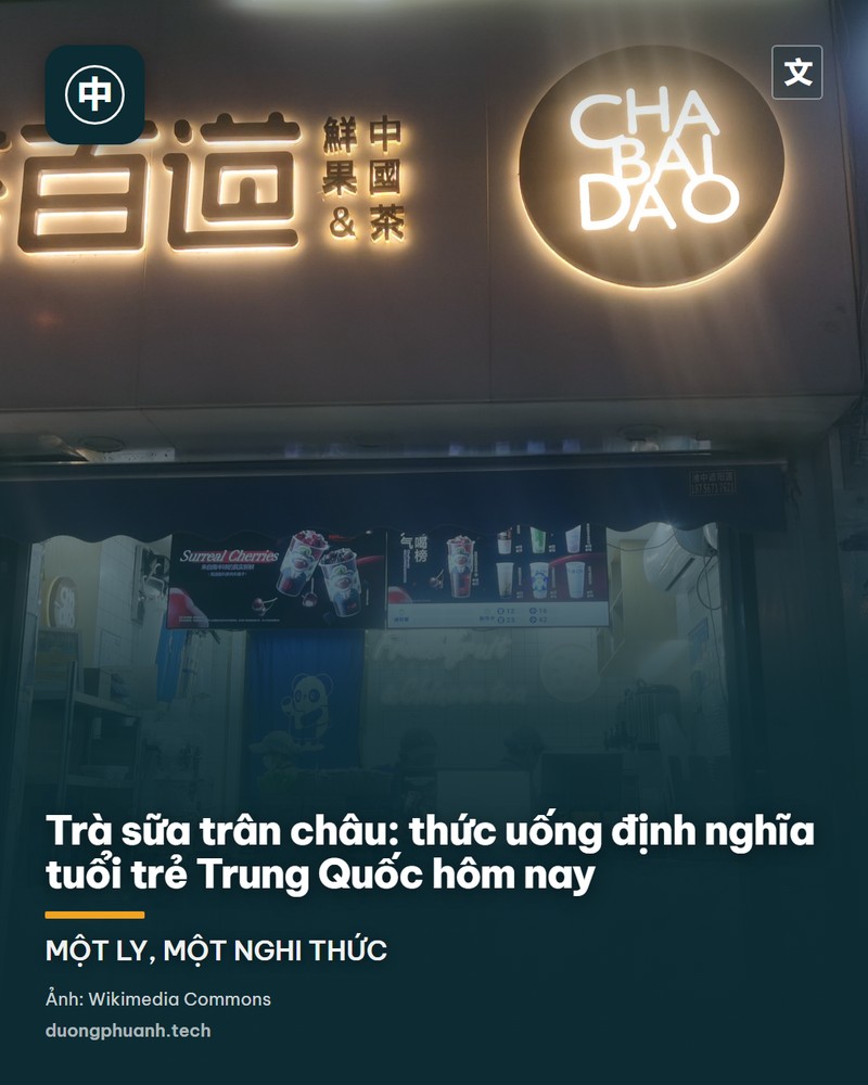 Một ly trà sữa trân châu cốt trụ trong ly thuỷ tinh cao, trên thân ly in logo chữ Hán màu đỏ, những hạt trân châu đen lắng dưới đáy, đặt trên bàn gỗ trong không gian quán ấm áp với ghế gỗ phía sau